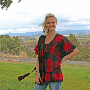 May include: A woman wearing a red and black plaid poncho with a black tank top underneath. She is standing in a field with a mountain range in the background.