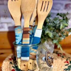 May include: A set of wooden kitchen utensils, including a spoon, spatula, and slotted spoon, with patterned fabric handles in shades of blue, brown, and cream. The utensils are displayed in a clear glass jar with a decorative lid. A woven placemat is beneath the jar.