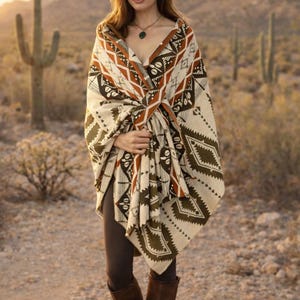May include: A woman wearing a cream-colored poncho with a geometric pattern in brown, black, and olive green. She is wearing dark leggings and brown leather boots. The background is a desert landscape with cacti and mountains.