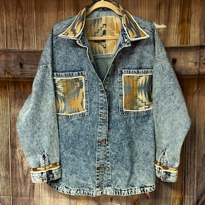 May include: A denim jacket with a light blue acid wash. The jacket features a patterned collar, pocket accents, and cuffs in shades of tan, brown, and cream. It has two front pockets and a button-down closure, hanging on a wooden hanger.