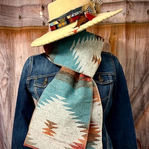 May include: A cream-colored fedora hat with a decorative band and feather accents sits atop a denim jacket. A turquoise, gray, and brown patterned scarf is draped around the jacket, creating a layered, southwestern-inspired look.