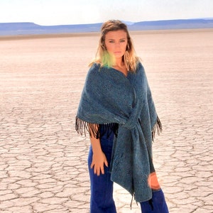 May include: A woman wearing a blue woven shawl with black fringe and blue jeans. The shawl is draped over the shoulders and tied at the waist. The background is a cracked, dry landscape under a bright sky.