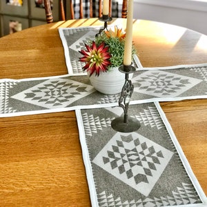 May include: A wooden table with a gray and white geometric patterned table runner. The table runner has a white border. A white ceramic vase with a green and red succulent plant is in the center of the table runner. Two white candles are in a black metal candlestick.