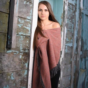 May include: A woman with long brown hair is wearing a pink and gray shawl with fringe. She is standing in front of a weathered wooden door.