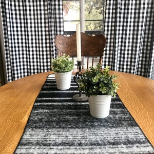 May include: Black and white checkered curtains frame a wooden table with a black and white striped table runner. Two white ceramic pots with green plants sit on the runner.