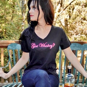 May include: A black V-neck t-shirt with the text "Got Whiskey?" in pink script. The image also shows a clear glass bottle of whiskey with a wooden stopper, and a shot glass. The person is sitting on a teal bench.