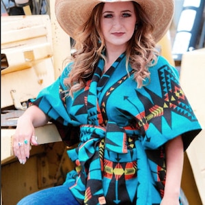 May include: A woman wearing a straw cowboy hat and a turquoise and black patterned wrap. The wrap is tied at the waist and features a geometric design in turquoise, black, yellow, and orange. She is also wearing blue jeans and turquoise jewelry.