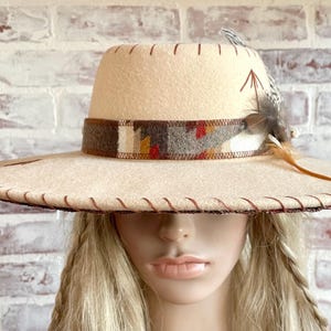 May include: A cream-colored fedora-style hat with a wide brim and a decorative band featuring a geometric pattern in brown, red, and yellow. The hat is adorned with feathers and stitched detailing.