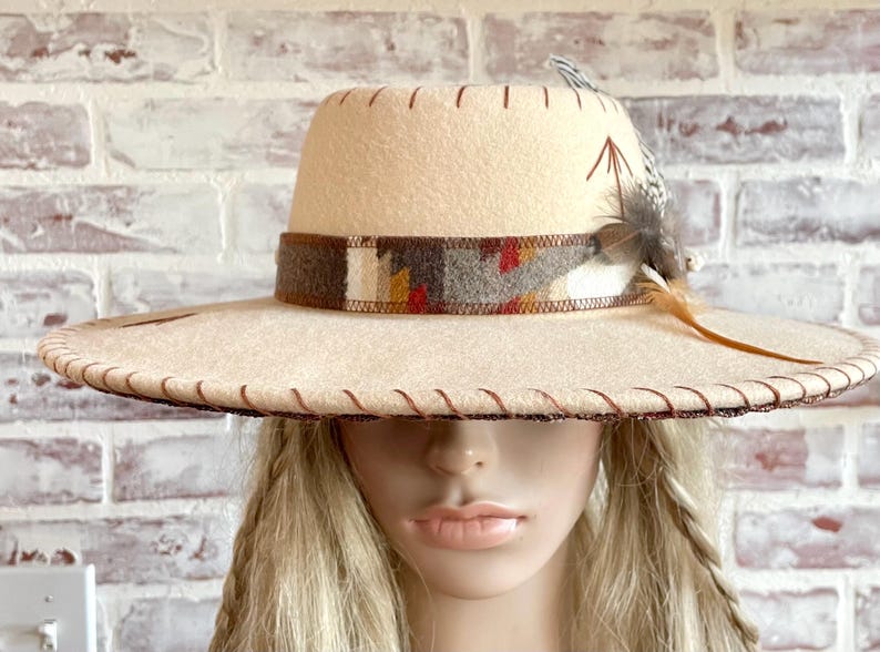 May include: A cream-colored fedora-style hat with a wide brim and a decorative band featuring a geometric pattern in brown, red, and yellow. The hat is adorned with feathers and stitched detailing.