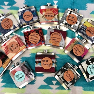 May include: A collection of 14 silver hip flasks with various colorful patterned fabric coverings and leather patches with different sayings. The sayings include "Get Whiskey", "HUYAMED COWGIRL", "DRINKING", "IF NECESSARY", "SIP SIP... Hooray!!", and "MAMA TRIED".
