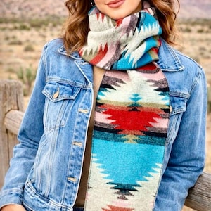 May include: A colorful patterned scarf with a turquoise, red, and cream design. The scarf is worn with a denim jacket and a cowboy hat. The scarf has a geometric pattern and is fringed at the ends.