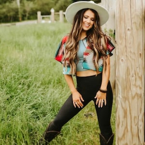 May include: A woman wearing a white hat, a colorful patterned crop top, black leggings, and brown boots. She is standing in a field of green grass.