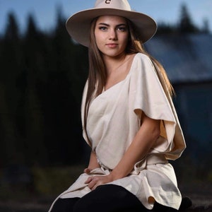 May include: A woman wearing a cream-colored, loose-fitting top with a wide neckline and short sleeves. She is sitting on a brown fur rug and wearing a wide-brimmed hat with a brown letter "C" on the front.