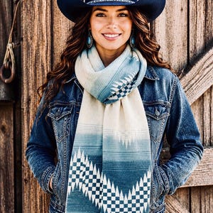 May include: A dark blue cowboy hat with a decorative band, a long scarf featuring a blue and cream gradient with a geometric triangular pattern, and a blue denim jacket. The scarf is wrapped around the neck and hangs down the front of the jacket.