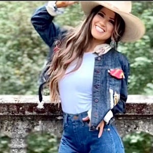 May include: A denim jacket with patches and a light-colored hat. The jacket has a white and floral design on the collar and cuffs. The person is wearing a white top and blue jeans.