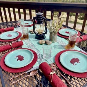 May include: A rustic outdoor dining table setting with turquoise plates featuring a bison design, set on red placemats. Includes a black lantern, glasses, silverware, and red napkins. The table is set on a metal mesh table with a scenic background.