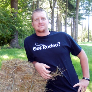 Puede incluir: Una camiseta azul marino con el texto "Got Rodeo?" en escritura blanca. La persona se apoya en una paca de heno en un entorno exterior cubierto de hierba con árboles en el fondo.