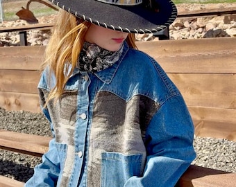 May include: A black cowboy hat with a white stitched brim and a patterned band. The hat is paired with a denim jacket featuring a patterned front panel and two front pockets. The jacket has silver buttons.