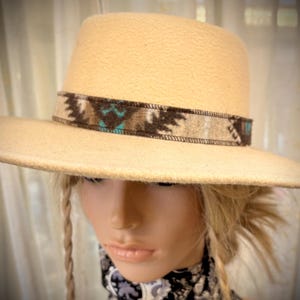 May include: A cream-colored fedora-style hat with a wide brim. The hat features a decorative band with a Southwestern-inspired pattern in shades of brown, beige, and turquoise. The hat is displayed on a mannequin head.