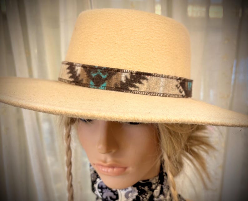 May include: A cream-colored fedora-style hat with a wide brim. The hat features a decorative band with a Southwestern-inspired pattern in shades of brown, beige, and turquoise. The hat is displayed on a mannequin head.
