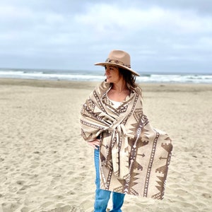 May include: A woman wearing a brown and white patterned shawl with a brown hat on a sandy beach.