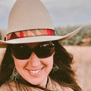 May include: A beige cowboy hat with a patterned band in red, brown, and orange. The hat is worn with sunglasses and a quilted vest. The image has a warm, outdoor aesthetic.