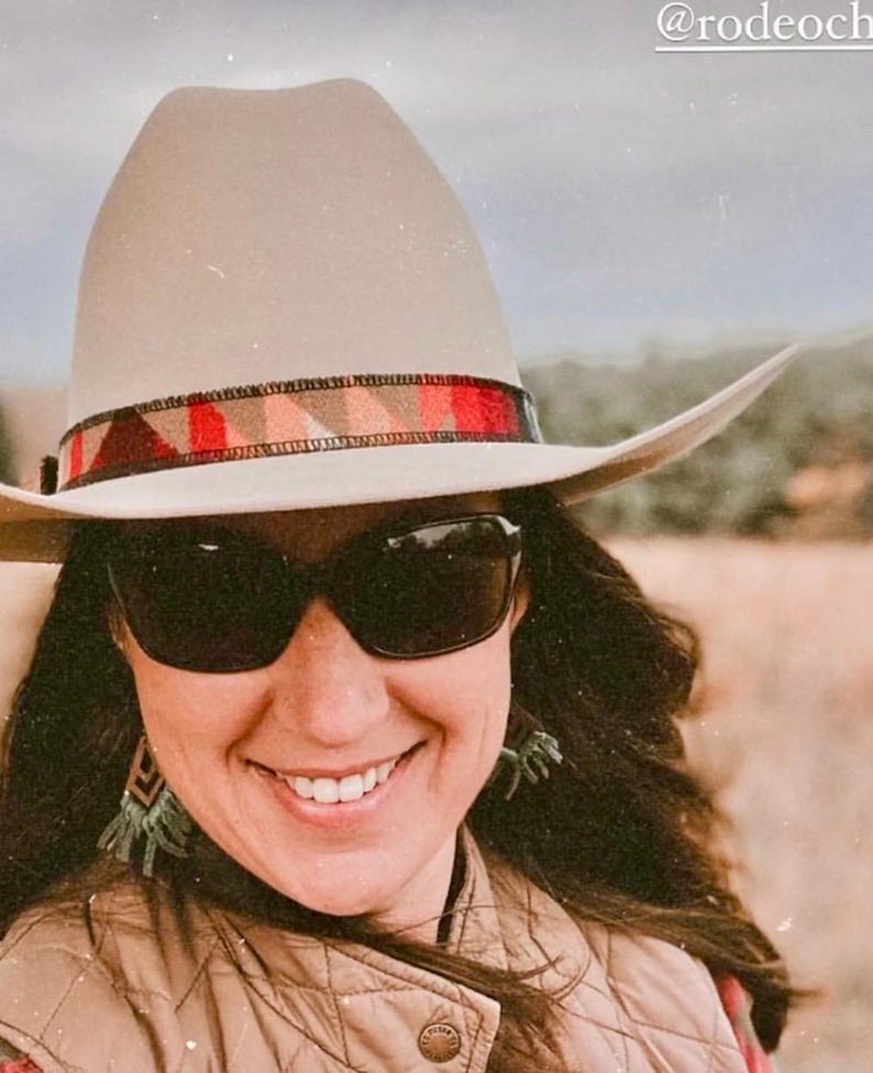 May include: A beige cowboy hat with a patterned band in red, brown, and orange. The hat is worn with sunglasses and a quilted vest. The image has a warm, outdoor aesthetic.
