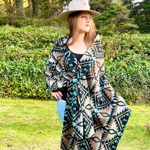 May include: A beige cowboy hat with a curved brim and a decorative emblem. A patterned shawl in shades of brown, black, and teal is draped over the shoulders. The shawl features a geometric design. The person is wearing blue jeans and a black top.