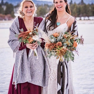 May include: Two women smiling, holding bouquets of flowers, and wearing shawls. One woman wears a burgundy dress and a gray shawl with fringe. The other wears a white lace dress and a patterned shawl. They are standing in a snowy landscape.