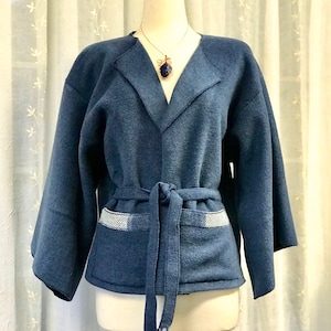 May include: A denim blue jacket with a shawl collar and a matching tie belt. The jacket has wide sleeves and pockets with a white and blue striped detail. A silver necklace with a blue stone pendant is also visible.