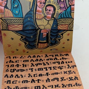 May include: Hand-painted religious icon featuring figures in blue, gold, and red. The artwork depicts a central figure with a halo, surrounded by other figures. The bottom portion displays text in a script. The overall style is reminiscent of traditional religious art.