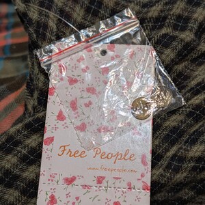 May include: A tag from Free People with a floral design and the brand's name in a script font. The tag is attached to a scarf with a plaid pattern in shades of brown, orange, and blue.