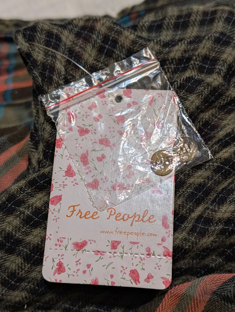 May include: A tag from Free People with a floral design and the brand's name in a script font. The tag is attached to a scarf with a plaid pattern in shades of brown, orange, and blue.