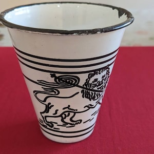 May include: White enamelware cup with a black and white illustration of a lion. The cup has a black rim and black stripes around the middle.