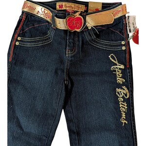 May include: Dark wash denim jeans with gold glitter "Apple Bottoms" script on the side. The jeans feature red stitching, a gold belt with an apple-shaped buckle, and decorative rivets. The belt has the words "Apple Bottoms" in red.