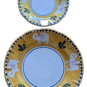 May include: Two round ceramic plates with scalloped edges, featuring a white center and a yellow rim decorated with white goat illustrations, green leaf motifs, and a blue dotted border. The plates have a smooth, glossy finish and a white base.