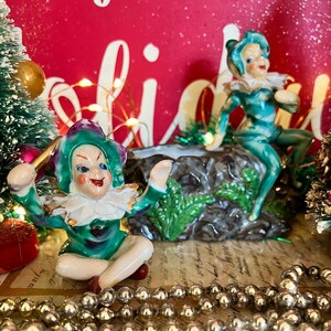 Könnte beinhalten: Zwei Keramik-Elf-Figuren mit grünen und lila Outfits, weißen Rüschen und rosigen Wangen. Einer sitzt im Schneidersitz, die Arme erhoben. Der andere liegt auf einer Felsformation. Silberne Perlenkette und Miniatur-Weihnachtsbäume im Hintergrund.