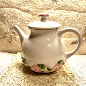 May include: White ceramic teapot with a pink lid and floral design. The teapot is sitting on a white crocheted doily.