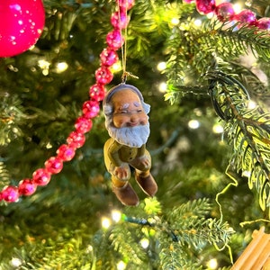 May include: A whimsical Christmas tree ornament depicting a smiling gnome with a white beard, wearing a green jacket and brown boots. The ornament is suspended from a gold string, surrounded by a Christmas tree with pink and silver beaded garland and a red ornament.