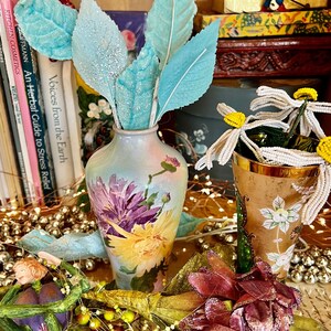 May include: A blue and white porcelain vase with floral designs sits on a table with a variety of decorative items, including a bouquet of blue and white leaves, a bouquet of white beads, and a pink and green flower.