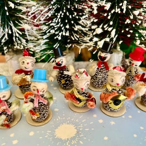 May include: A collection of nine vintage pine cone snowman ornaments, each with a different facial expression and wearing a hat. The ornaments are decorated with colourful felt and glitter, and some are holding musical instruments.