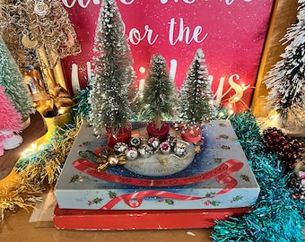 May include: A vintage Christmas gift box with a blue and white design featuring a snowy scene and the text "Christmas Greetings 24 Greeting Cards". The box is decorated with a red ribbon and holly sprigs. The box is surrounded by miniature flocked Christmas trees and red and gold tinsel.