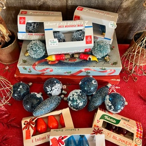 May include: A selection of vintage Christmas light bulbs in boxes. The bulbs are frosted and have a snowy appearance. Some are red, some are blue, and some are clear. The boxes are labelled with the brand name and the type of bulb. The boxes are on a red tablecloth.