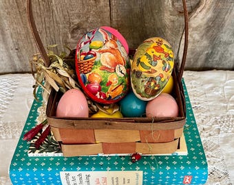 May include: A wicker basket filled with colourful Easter eggs, including a pink egg, a yellow egg, a blue egg, and two decorated paper eggs. The basket is sitting on top of a blue book with white text.