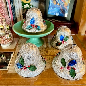 Pode incluir: Quatro enfeites de sino prateados com superfície texturizada, decorados com detalhes florais azuis, vermelhos e verdes. Um sino está sobre um pedestal verde, os outros sobre uma superfície de madeira. Os sinos são festivos e decorativos.