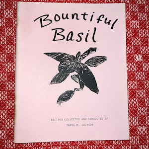 May include: A pink booklet with the title "Bountiful Basil" in black script. The cover features a black and white illustration of a basil plant. The text "Recipes collected and concocted by Tanya M. Jackson" is printed in black at the bottom of the cover.