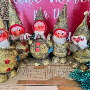May include: Five vintage Christmas gnomes made of paper mache with glitter and red accents. The gnomes are wearing hats and have white beards. They are all standing on a table with a piece of paper underneath them.