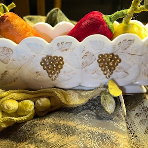 May include: A white ceramic bowl with a grapevine design and gold accents. The bowl is filled with fabric fruit, including a red pepper, a yellow pear, and an orange carrot.