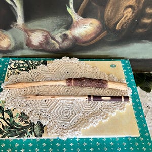 May include: Two wooden wands with brown and white accents, lying on a white crocheted doily on top of a book with a green cover.