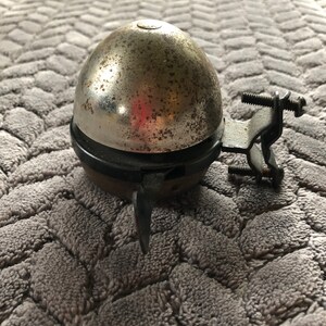 May include: A vintage silver bicycle bell with a black base and a metal bracket. The bell has a worn, aged look with rust spots.